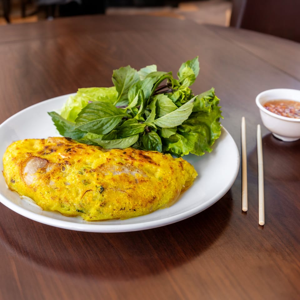 Vietnamese Banh Xeo crepe with lettuce, herbs, dipping sauce, and chopsticks on a wooden table.