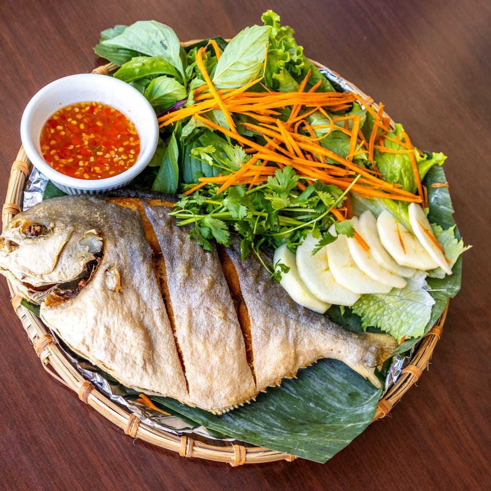 A whole fried fish with herbs, shredded carrots, sliced apples, and chili dipping sauce on a bamboo tray.