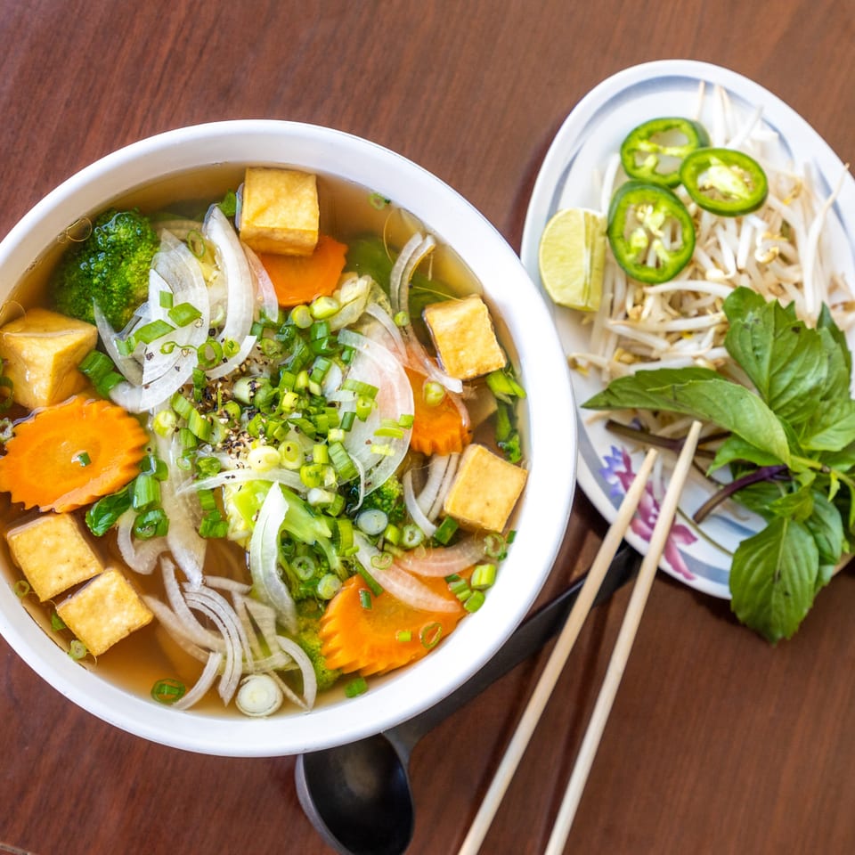 Vegetarian pho with tofu, broccoli, carrots, and onions, served with a side of fresh herbs.